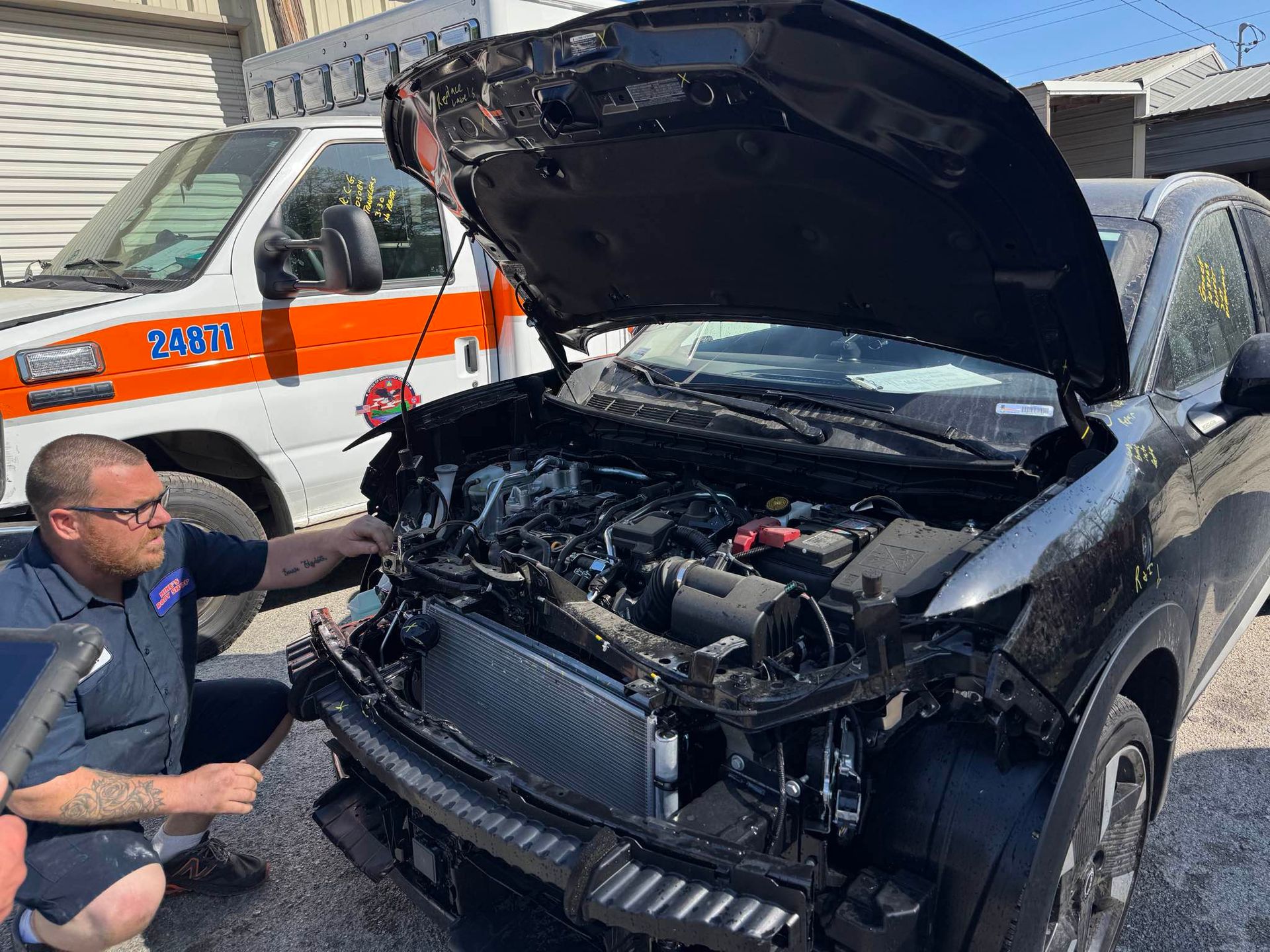 A mechanic examines the open engine bay of a black vehicle, with an orange-and-white emergency truck parked behind it.