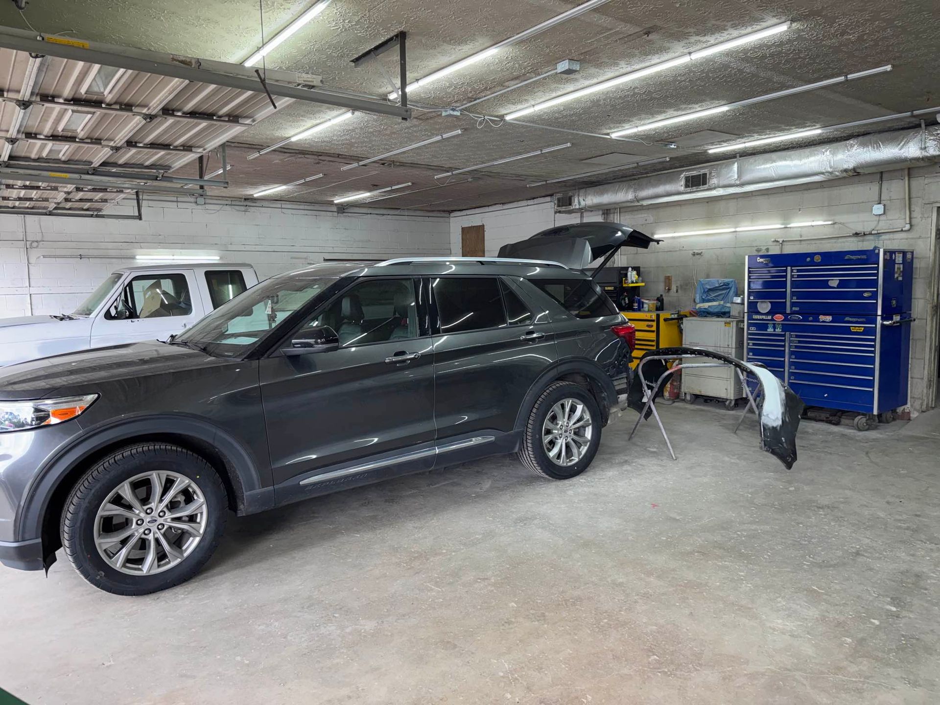 A dark gray SUV parked in an auto repair shop with a detached bumper on a stand near a blue toolbox.