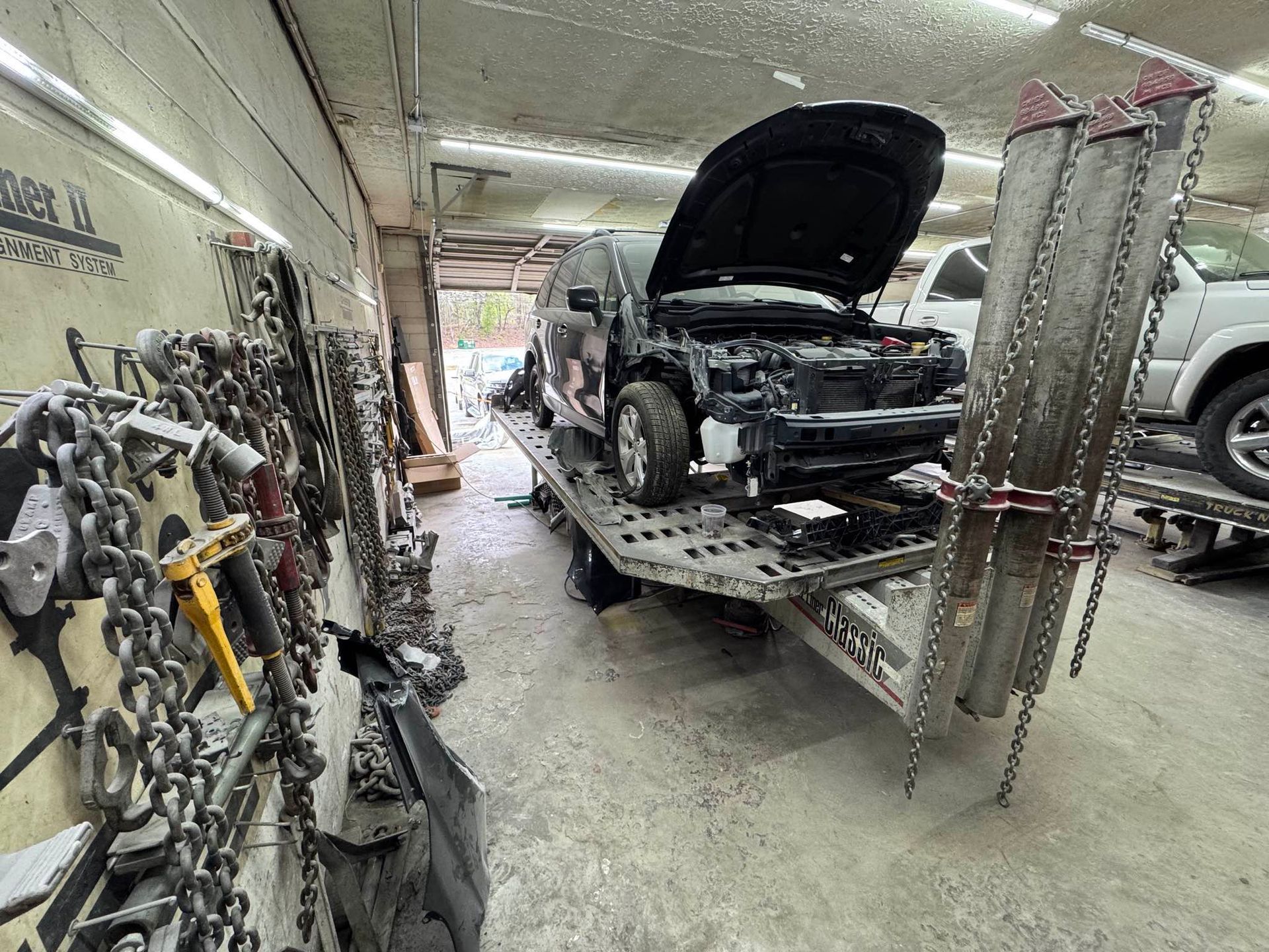 A dark damaged vehicle sits on a flatbed trailer inside a garage, with chains hanging on the wall to the left.