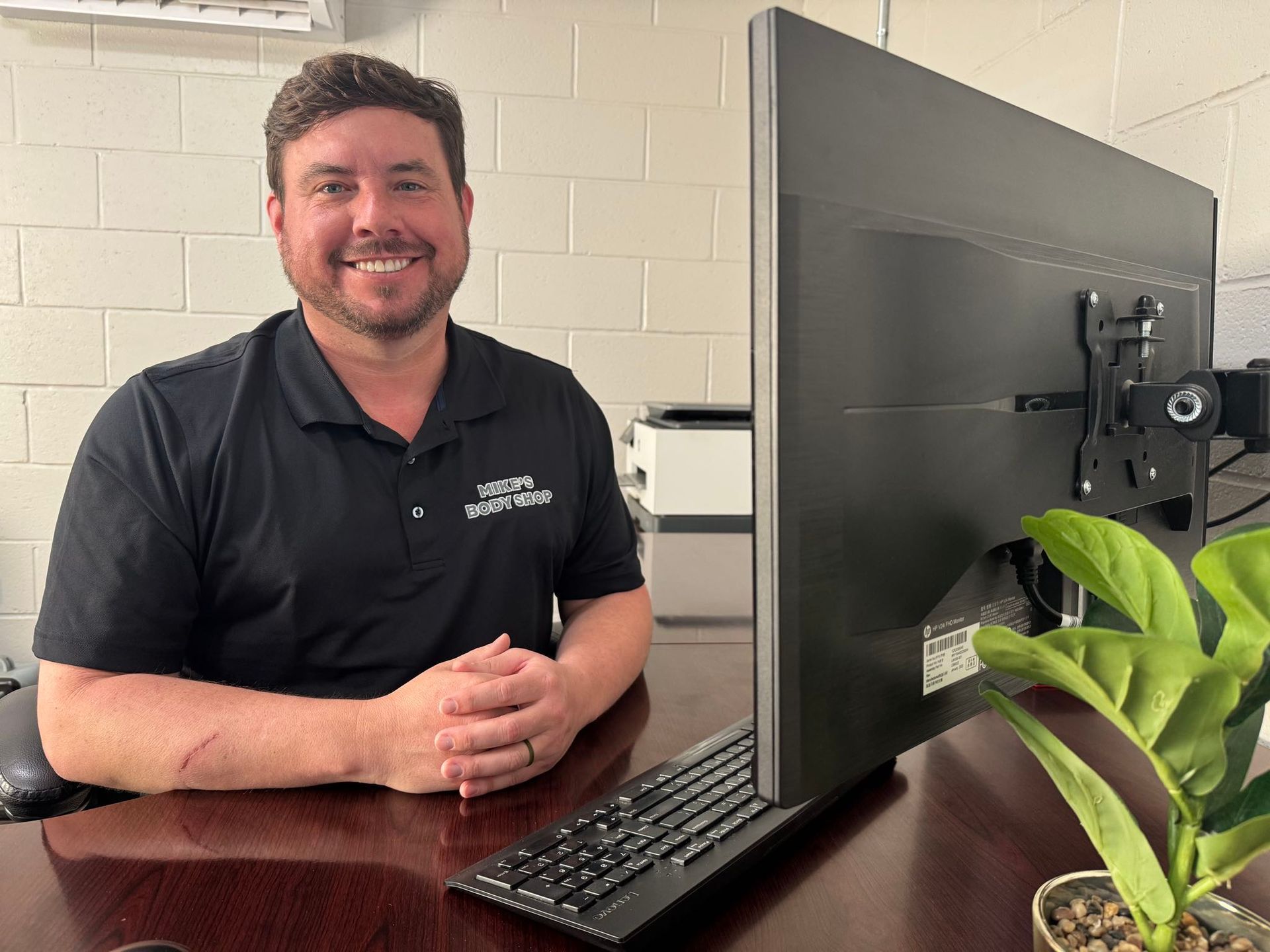 A smiling person in a black polo shirt sits at a desk with a computer monitor and a small potted plant in an office.