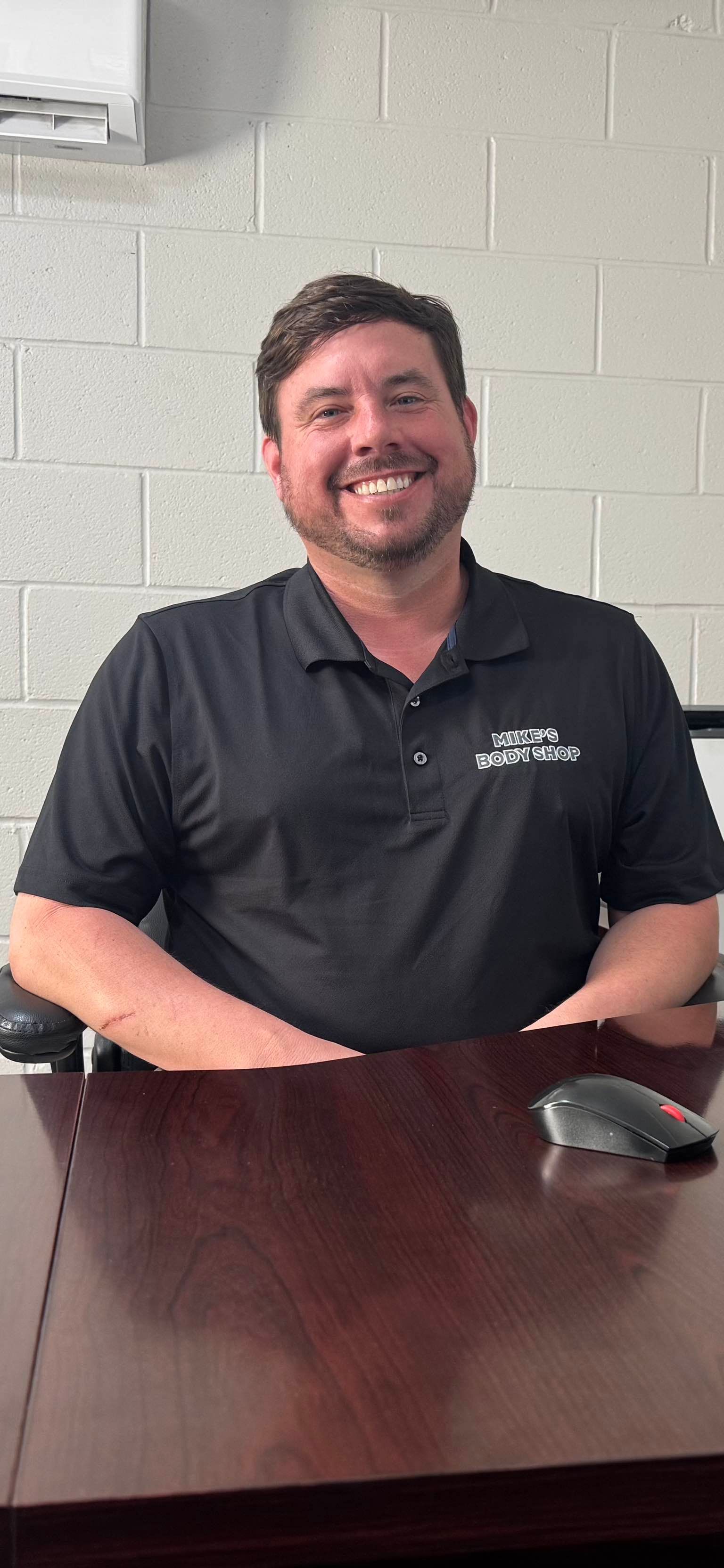 A smiling person in a black polo shirt with a logo sits at a dark wooden desk in front of a white cinder block wall.