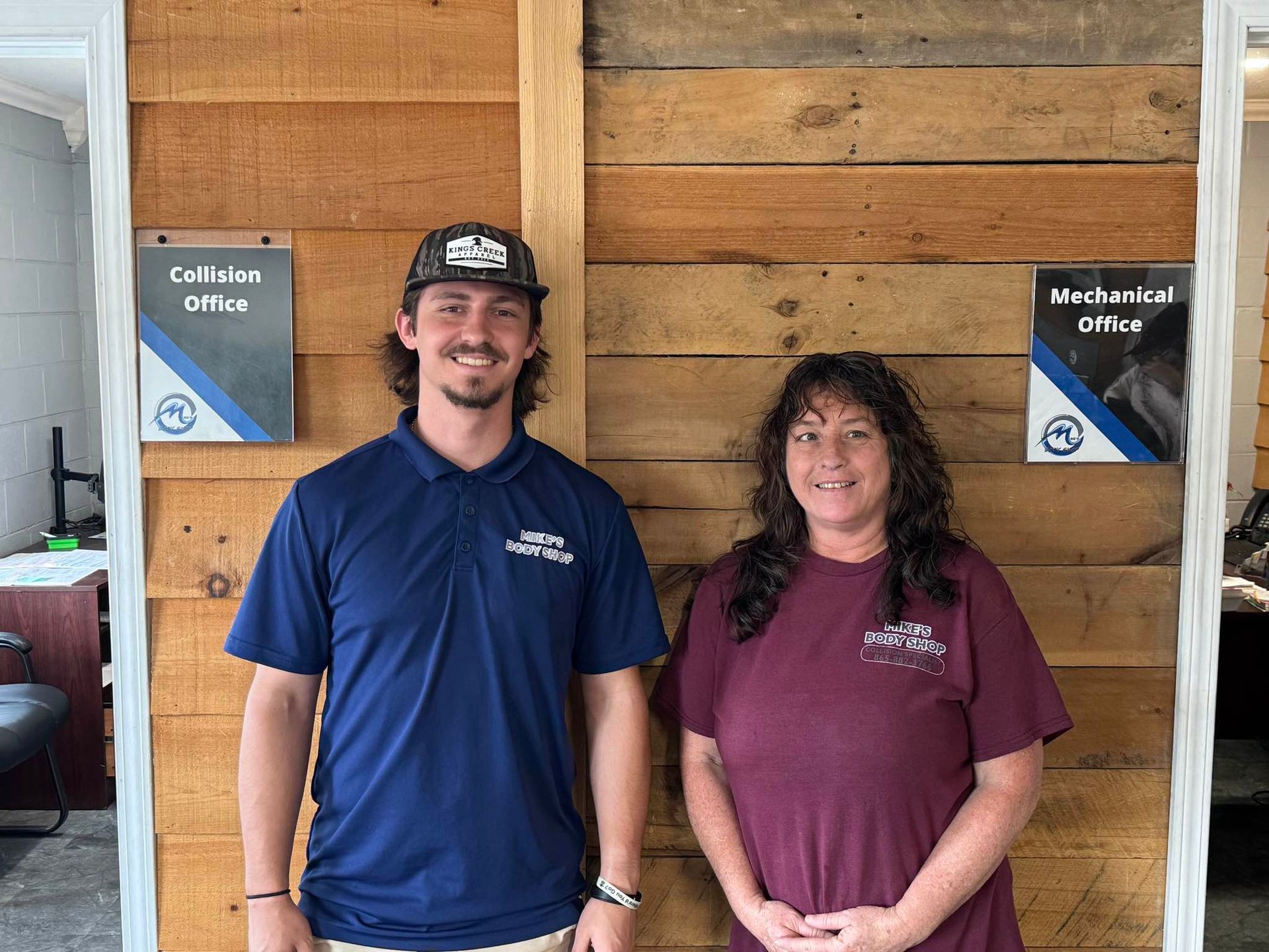A person in a blue polo and a person in a maroon shirt stand side-by-side in front of a wood-paneled office wall.