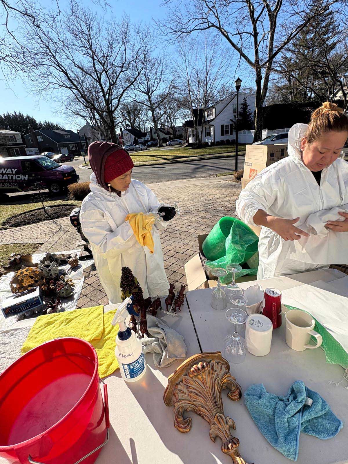 Two people in white protective suits clean household items on a folding table outdoors on a sunny day.
