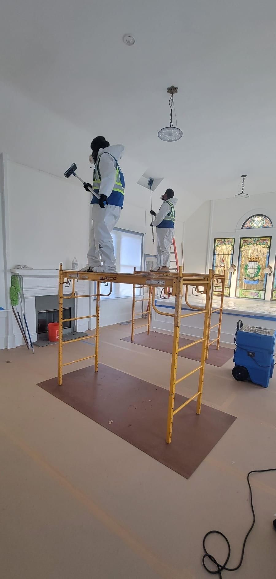 Workers painting a room from scaffold towers, with white walls and a brown floor covering