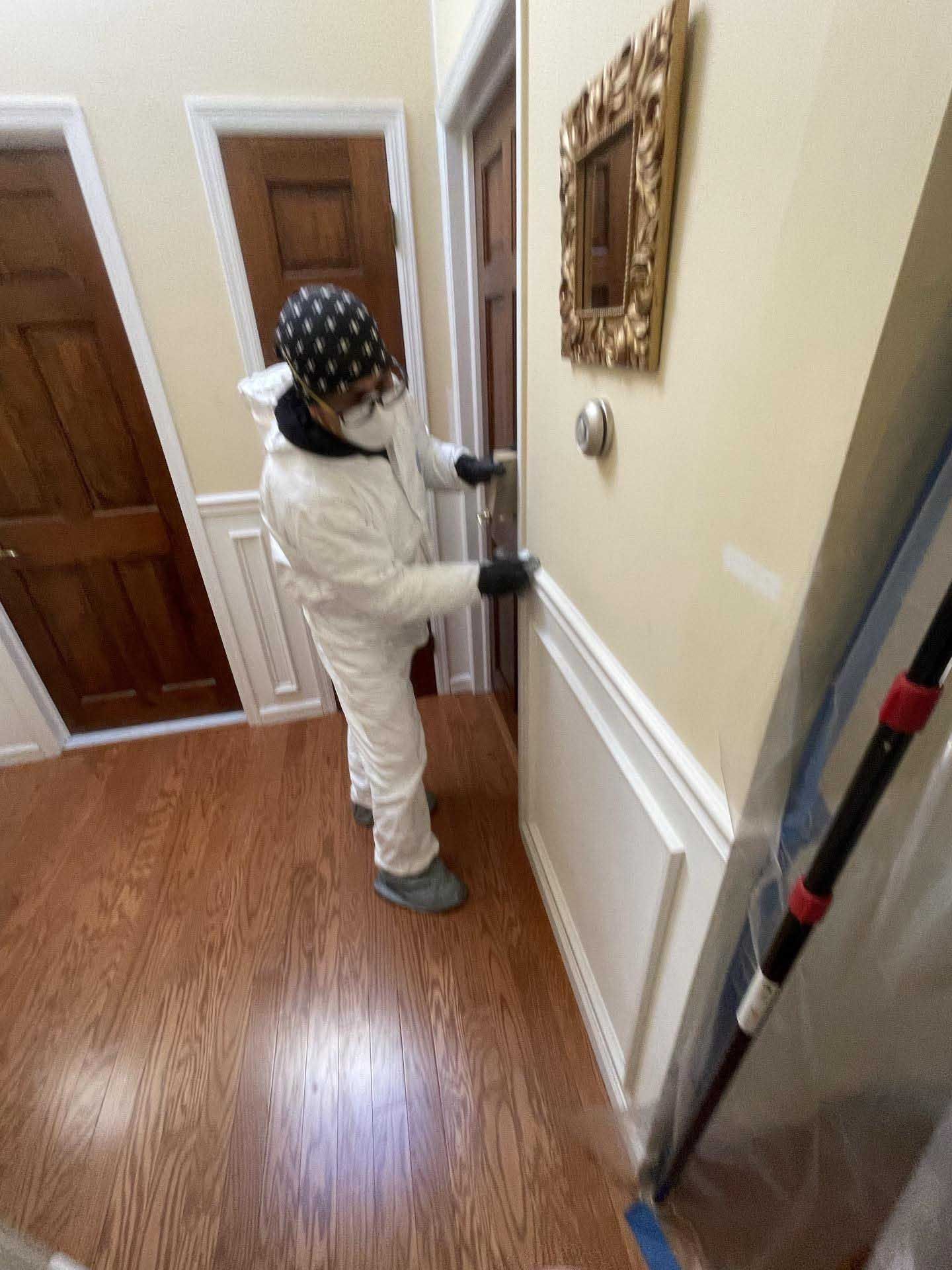 A person in white protective gear and a patterned head covering cleans a door handle in a residential hallway.