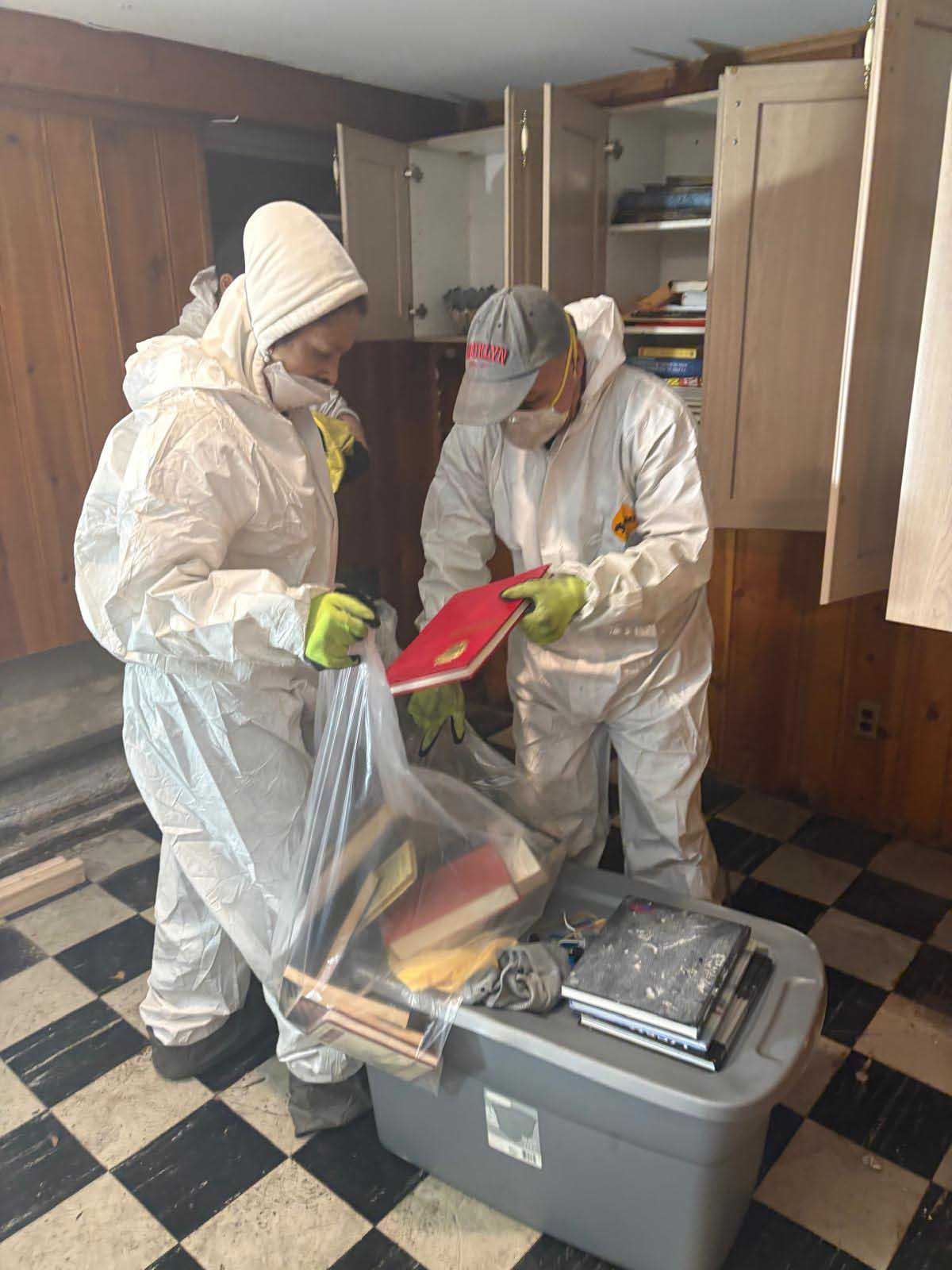 Two people in white protective suits and masks packing books into a plastic bin in a room with black-and-white tile floors.