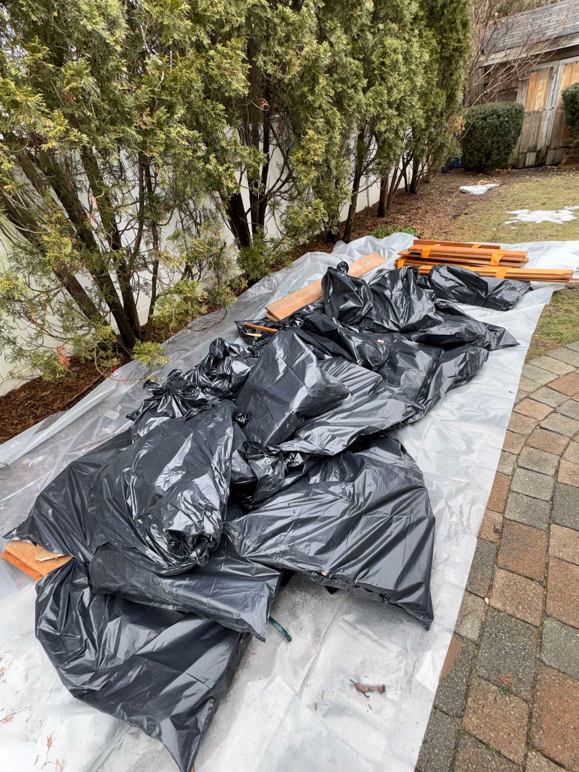 A pile of black trash bags sits on a white tarp on a paved area next to green trees and some scattered wooden planks.