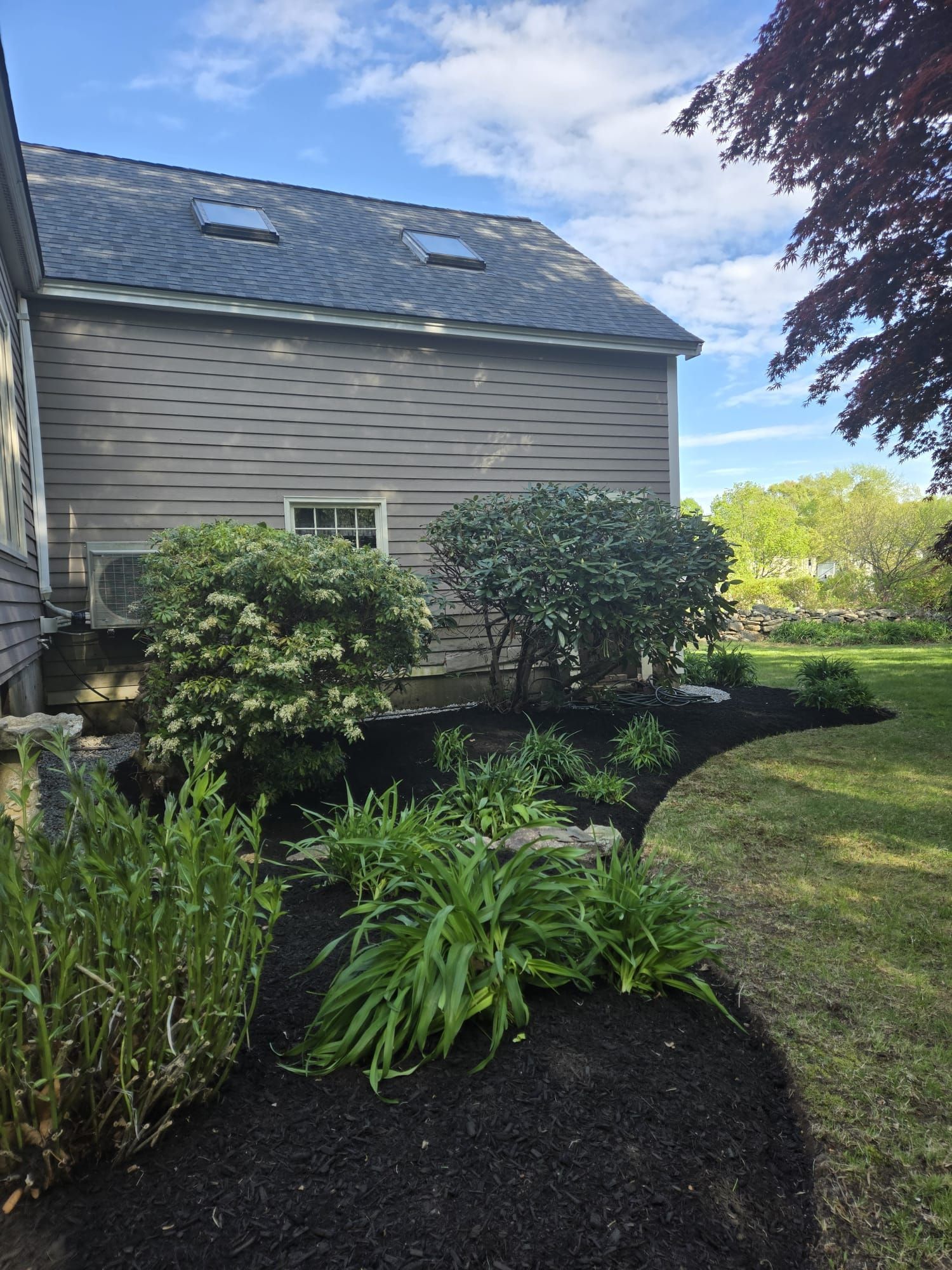 A landscaped garden bed with dark mulch, shrubs, and perennials curves along the side of a grey house under a blue sky.
