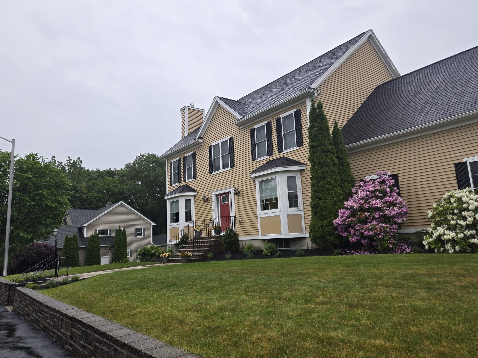 A yellow two-story house with a grey roof, bay windows, and a landscaped yard under a cloudy sky.