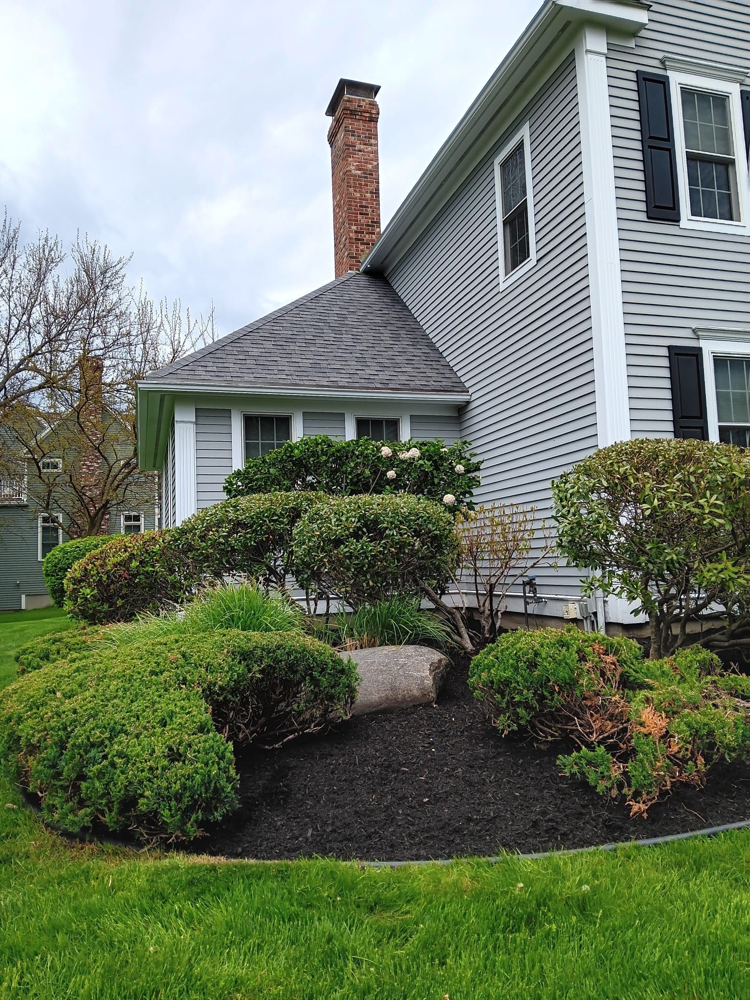 A grey house with a brick chimney and dark shutters sits behind a manicured garden with a large central boulder.