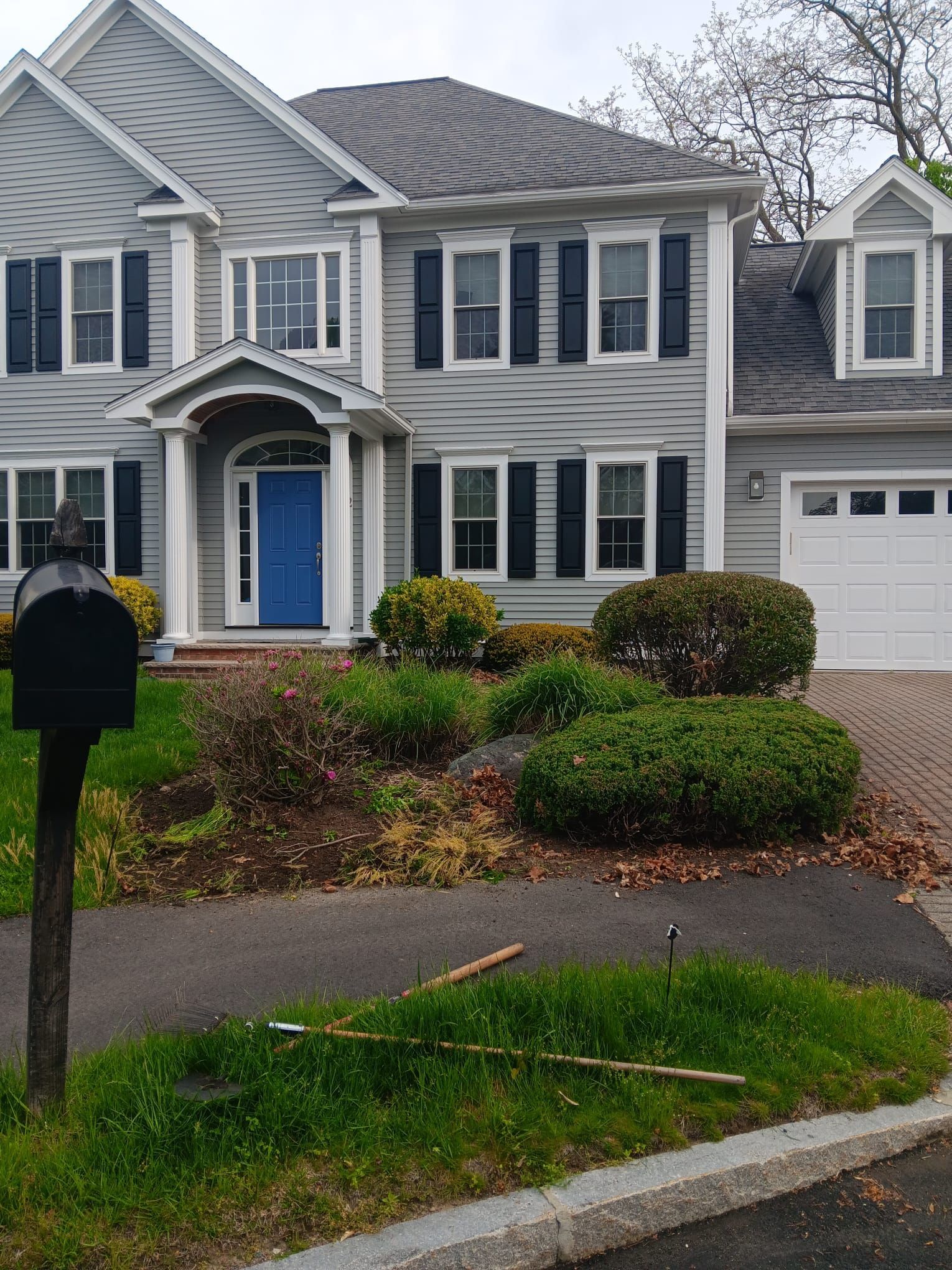 A two-story grey suburban house with a blue front door, white trim, black shutters, and front yard landscaping.