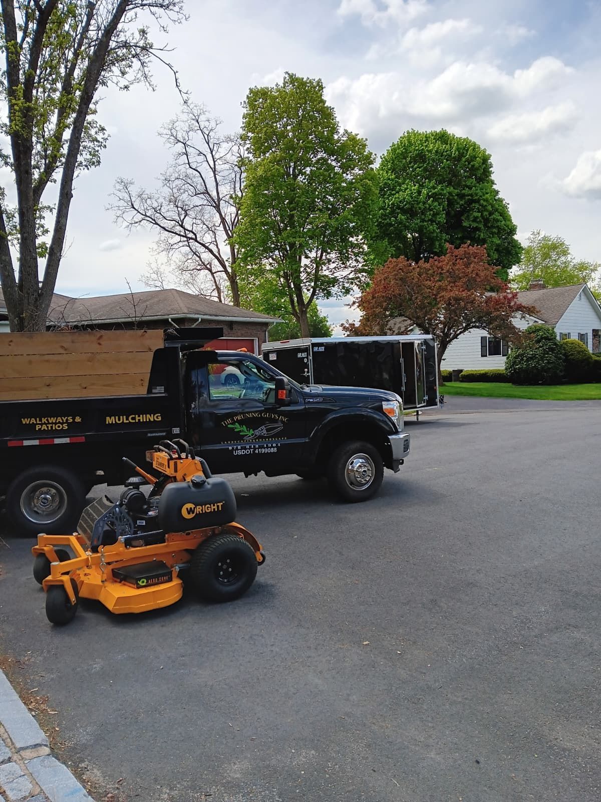 A yellow zero-turn lawn mower sits on an asphalt driveway next to a black utility truck and a trailer.