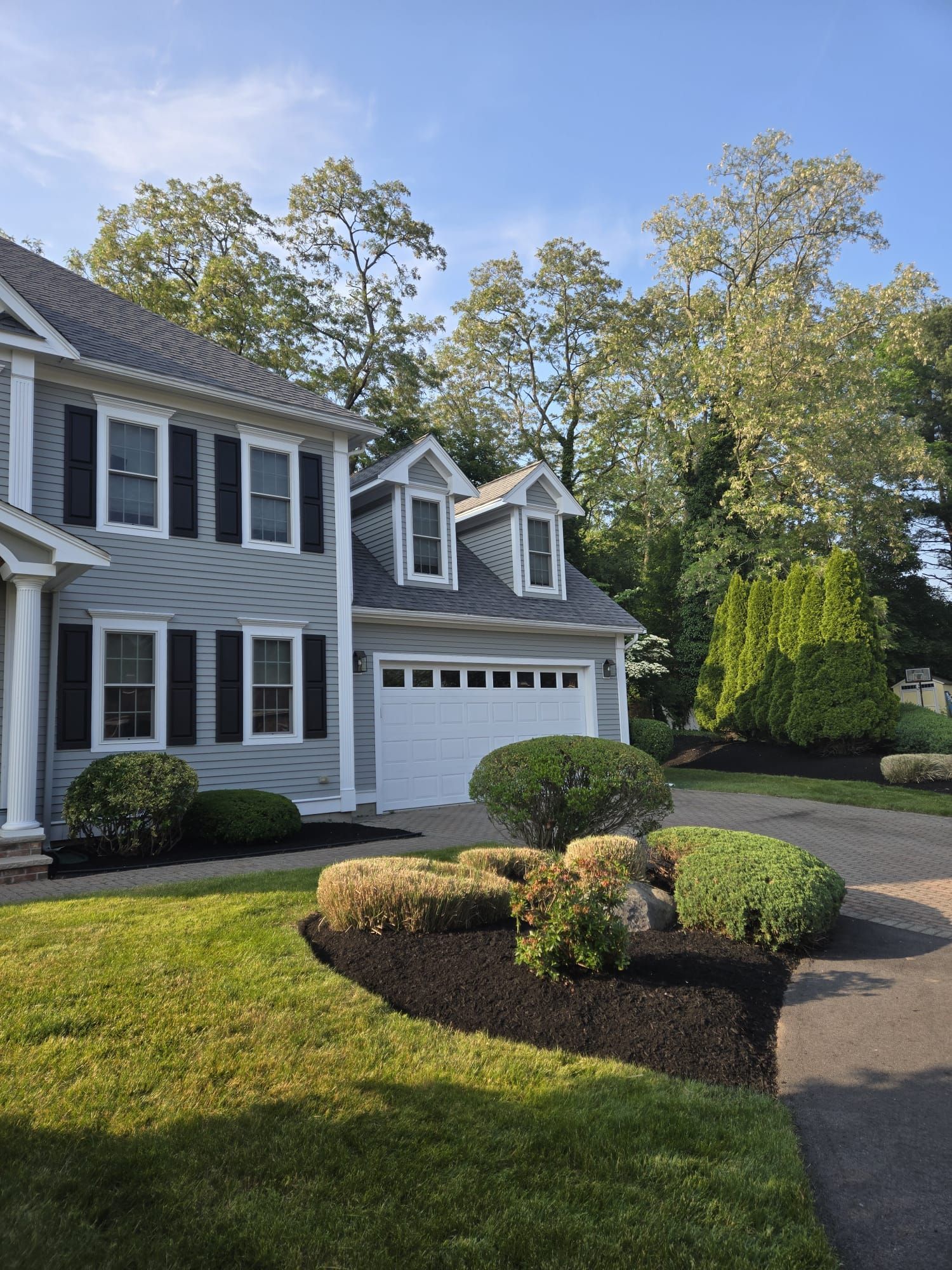A light gray two-story house with black shutters, a white garage, and a landscaped front yard with green bushes and mulch.