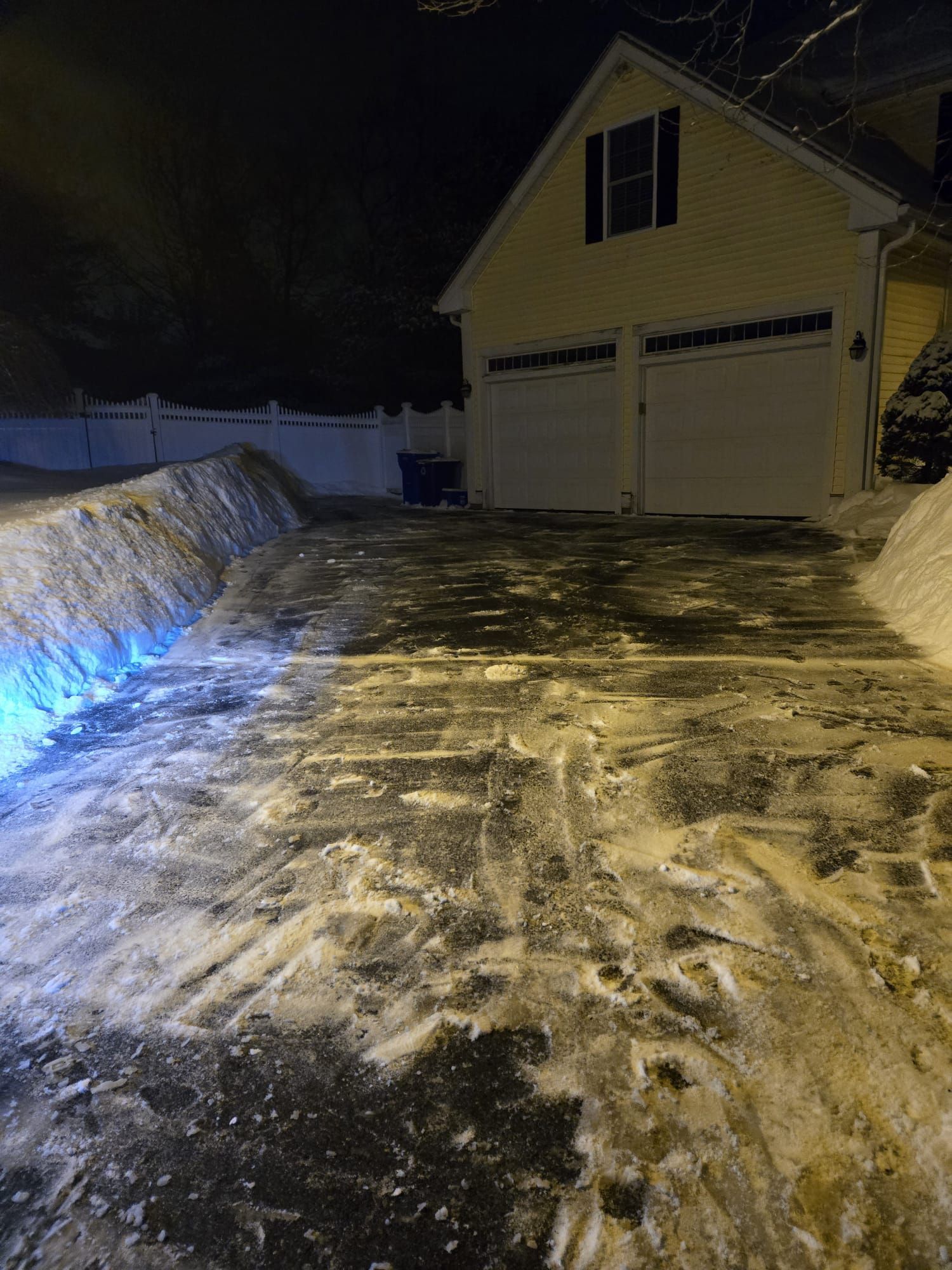 A yellow house with a two-car garage at night, overlooking a snow-covered, icy driveway cleared of deep snow banks.