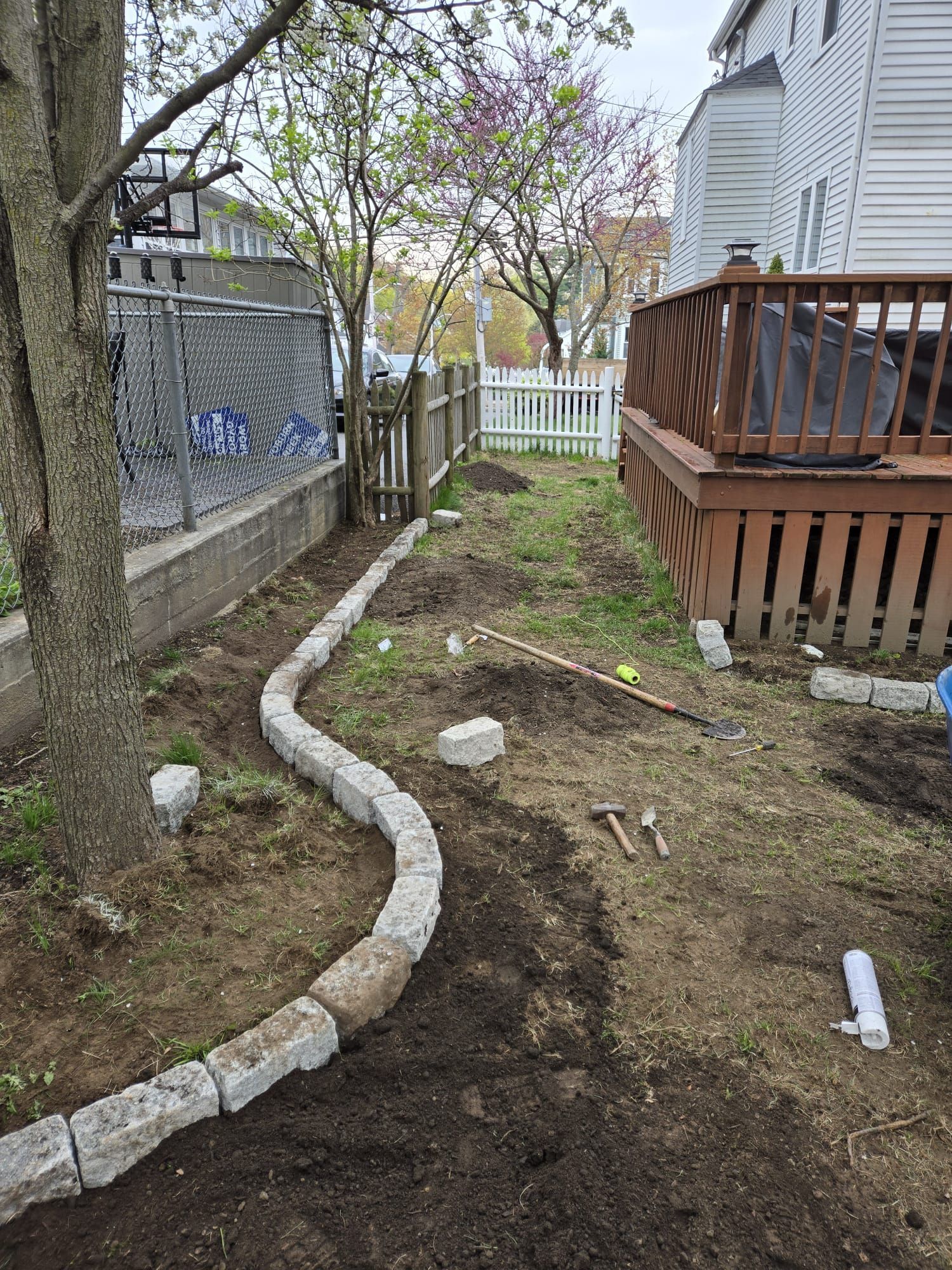 A curved stone garden border being installed in a backyard next to a tree, a wooden deck, and a white picket fence.