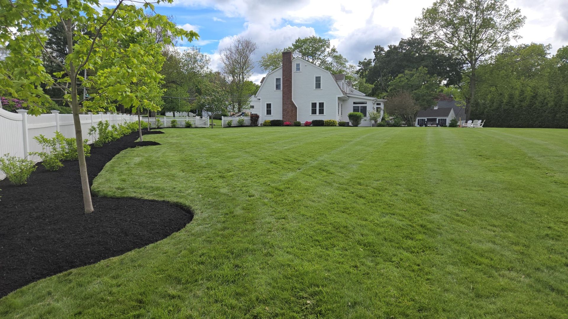 A white house with a chimney stands in the background behind a large, green lawn with a curved black mulch garden bed.