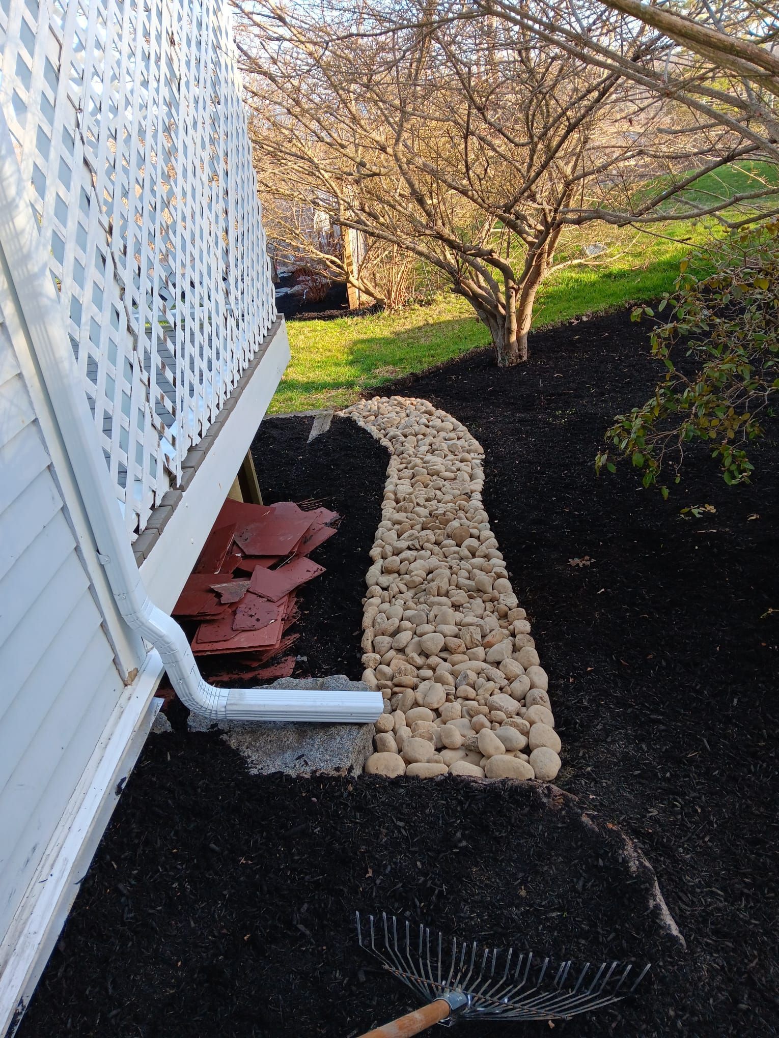 A garden path made of tan river stones winds along a white house foundation next to a downspout and dark mulch.