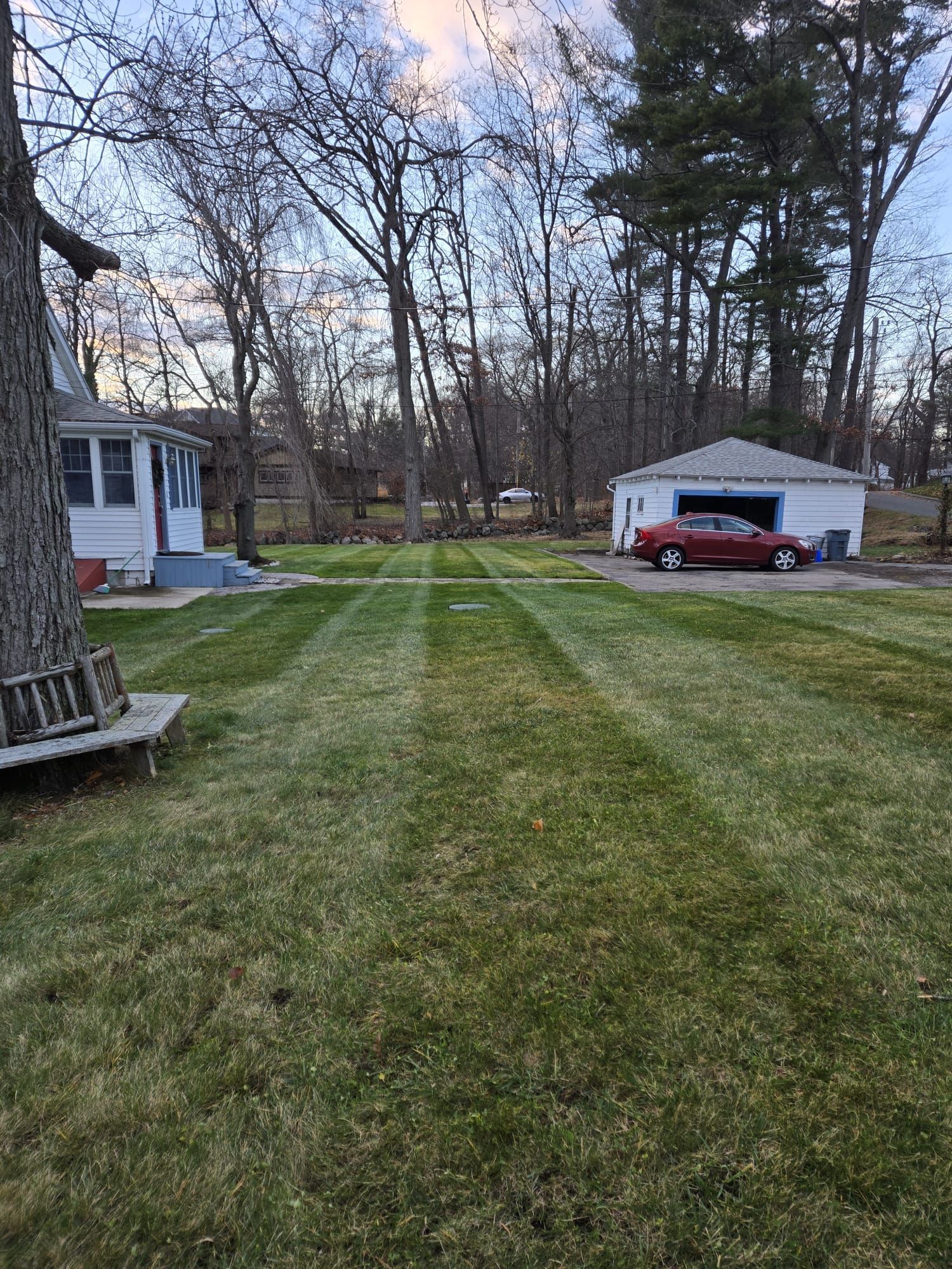 A large, freshly mowed lawn with distinct stripes stretches between a white house and a detached garage with a red car.