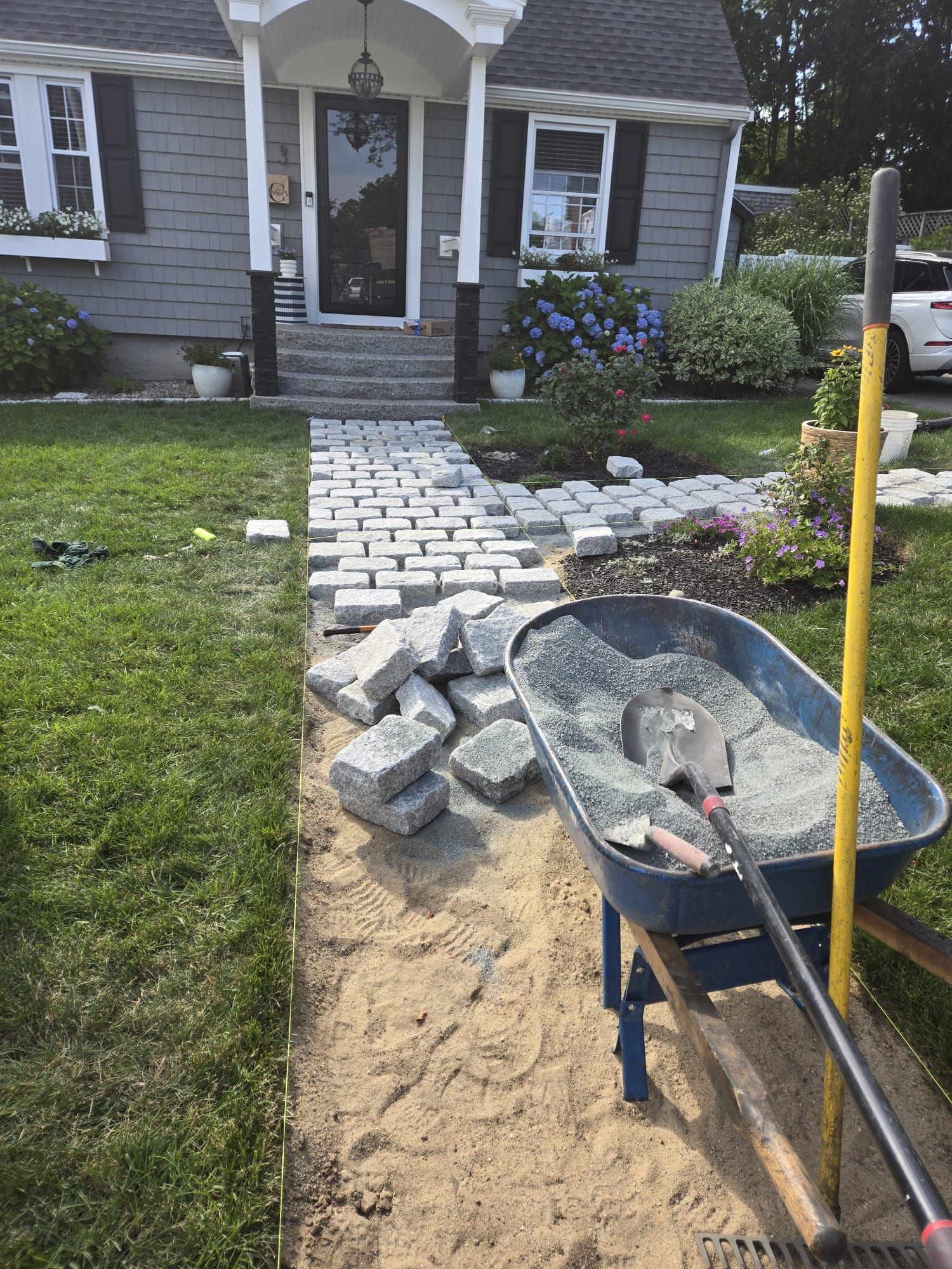 A partially installed stone walkway leading to a grey house, with a wheelbarrow of gravel and tools in the foreground.