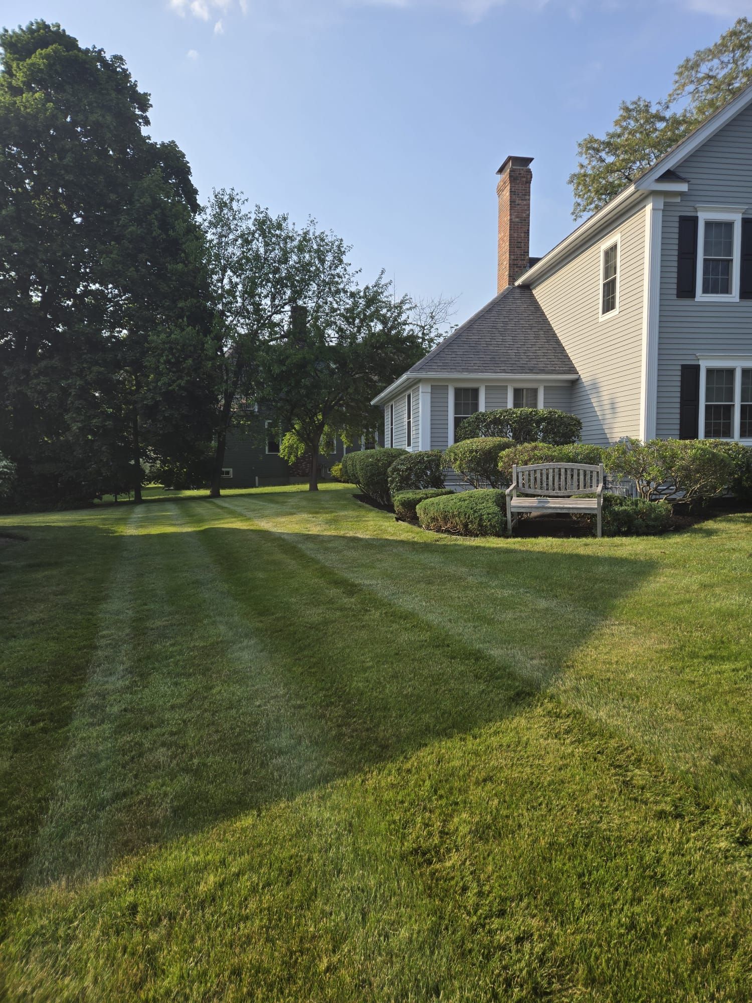 A house with gray siding and a chimney next to a large lawn with freshly mowed grass patterns and a wooden bench.