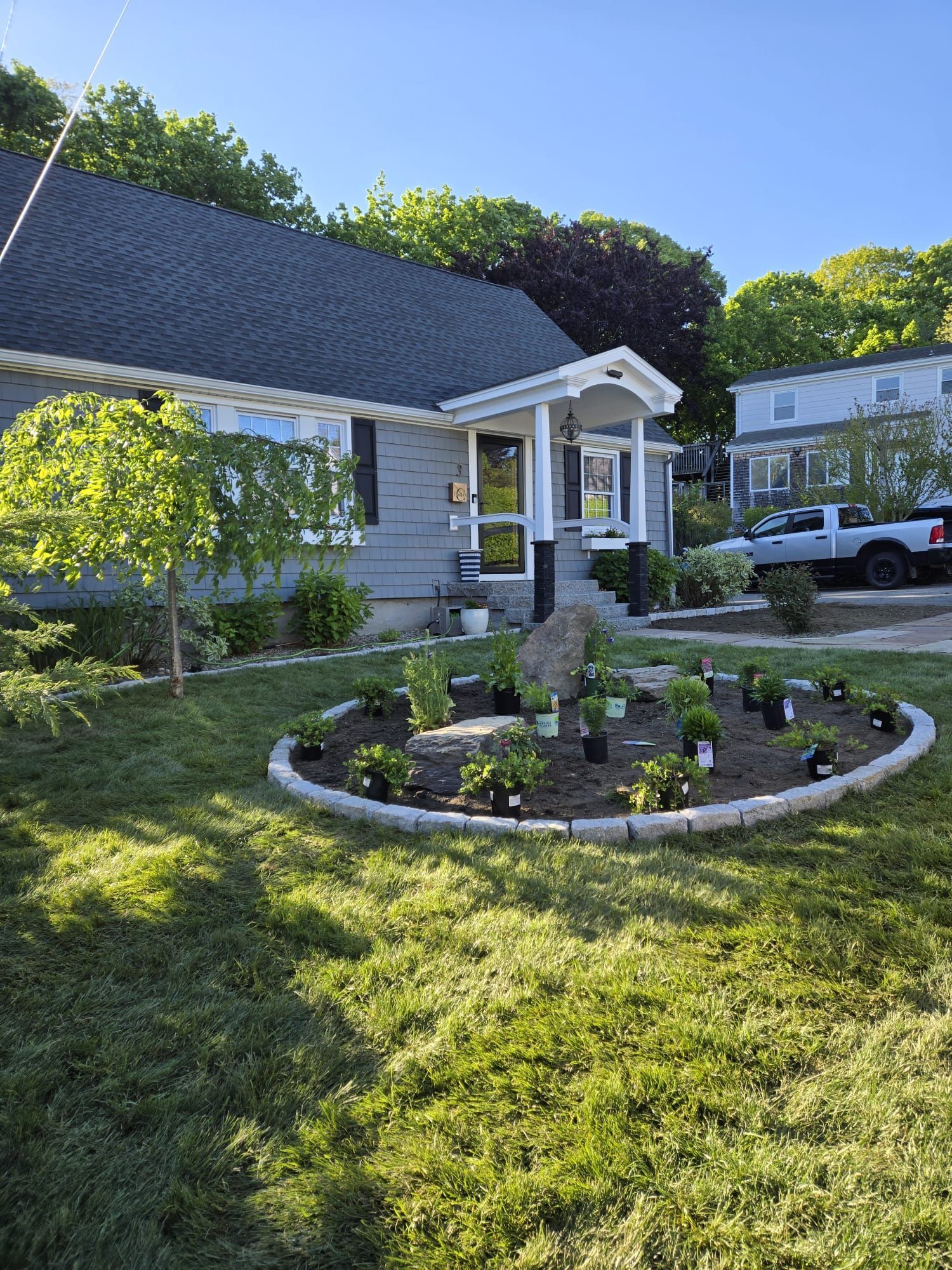 A gray house with a stone-bordered garden bed in the front yard featuring small plants and a decorative rock.
