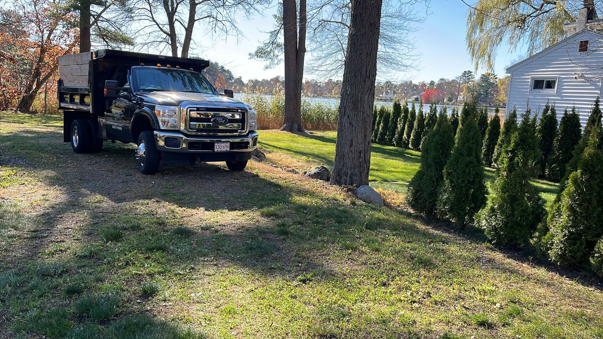 A dark-colored dump truck parked on a grassy lot next to a line of evergreen trees and a small white building.