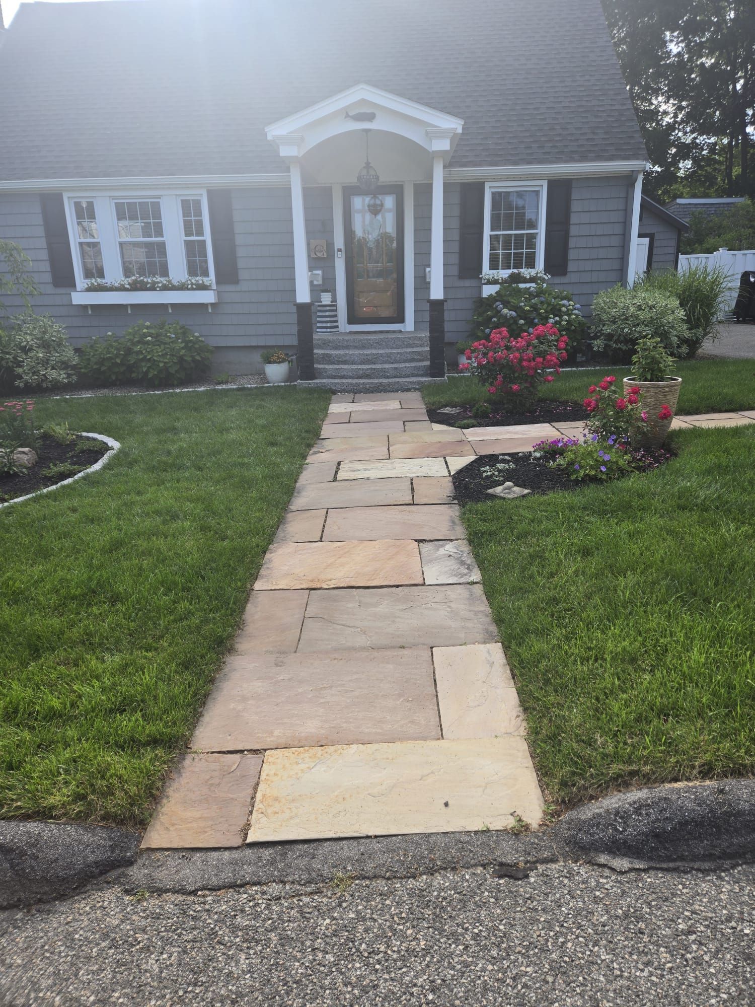 A stone walkway leads to the front door of a gray house with a white-trimmed entryway and landscaped front yard.