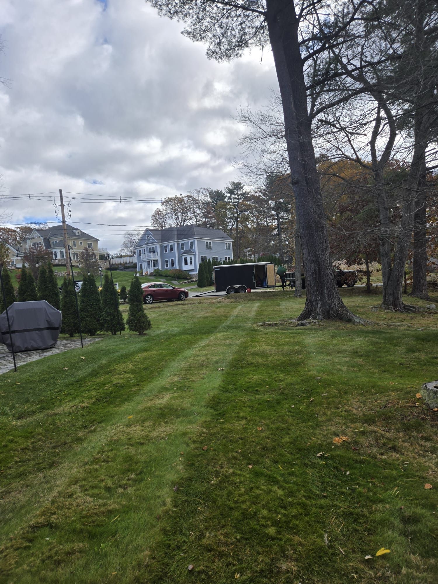 A mowed lawn leads toward a gray house and trees under a cloudy sky, with a dark trailer and a red car parked nearby.