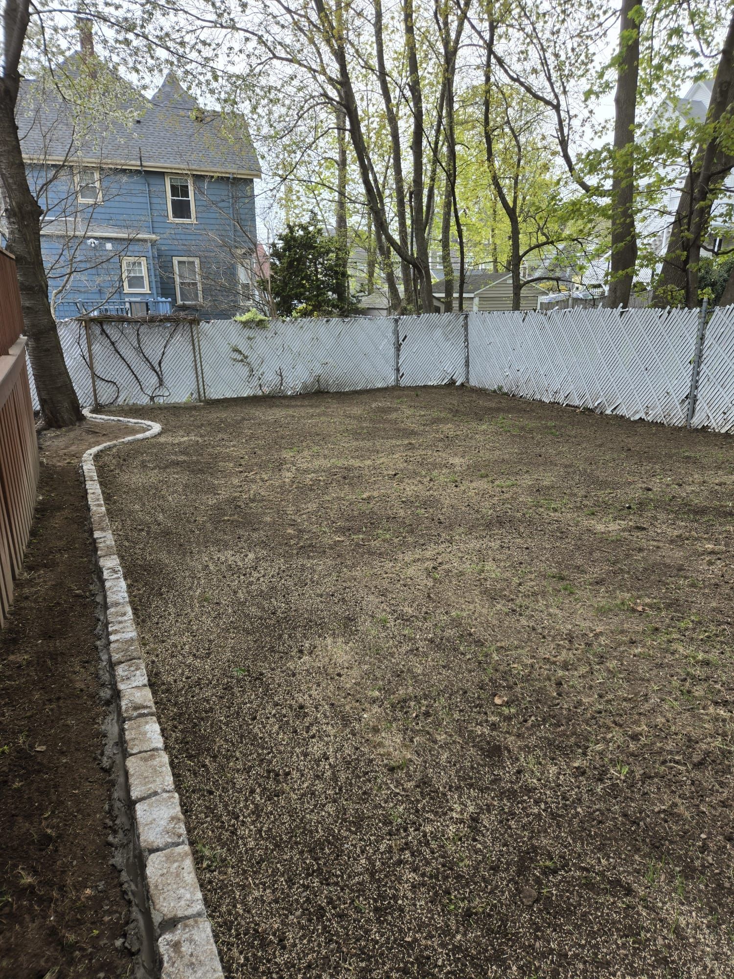 A backyard with a stone-bordered dirt lawn, a white slatted fence, and a blue house in the background.