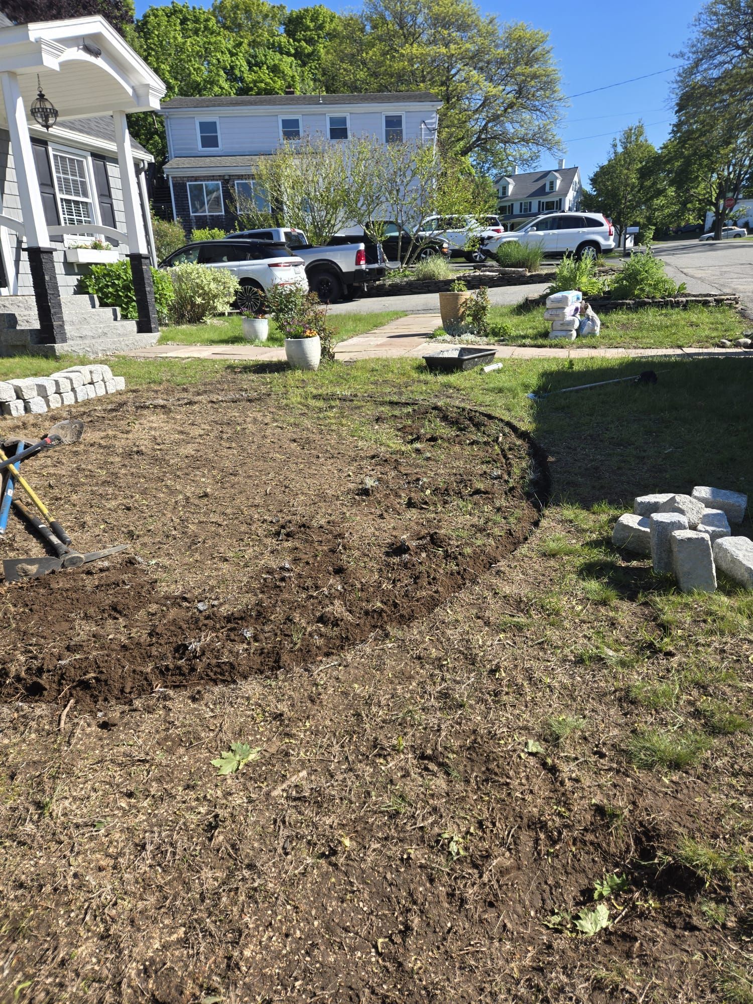 A yard with a newly dug, curved soil bed and stacks of concrete landscaping blocks near a house.