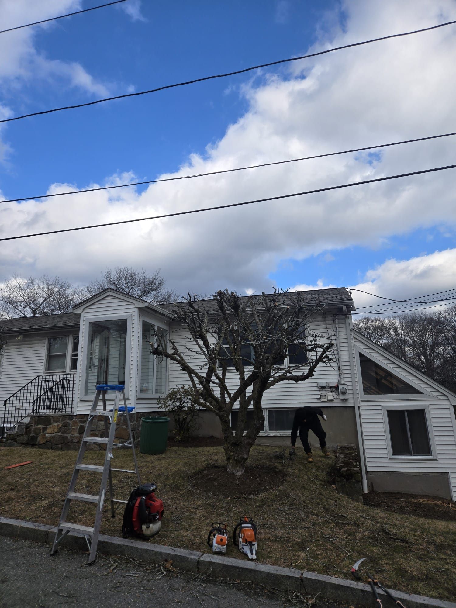 A person works on landscaping in front of a white house with tools, a ladder, and power saws on the lawn.
