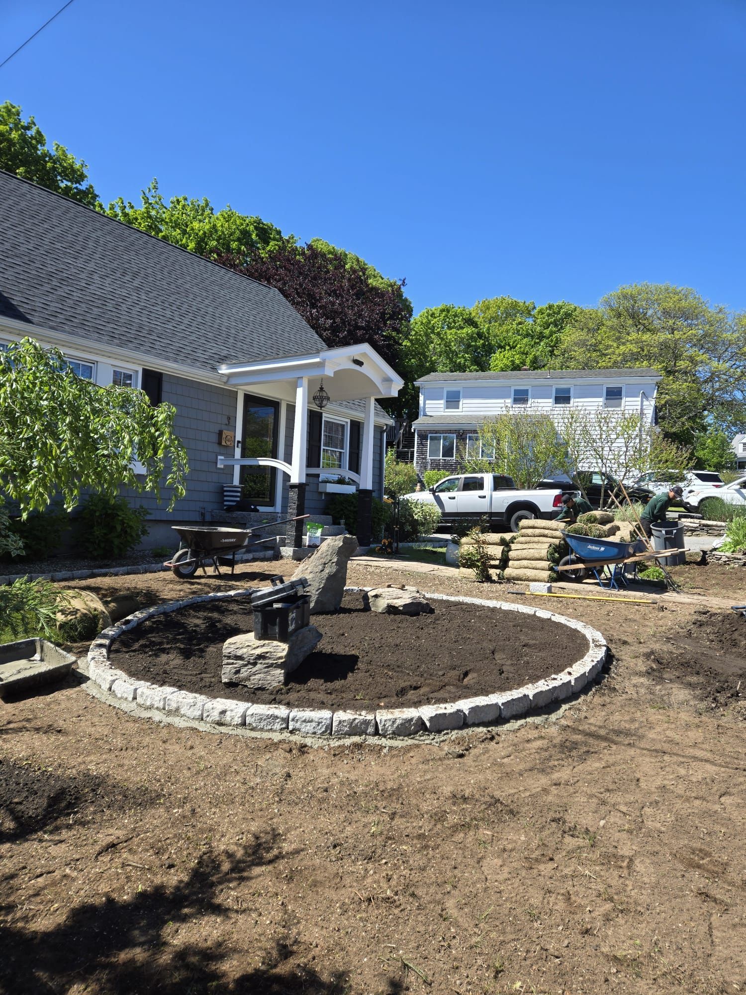 A front yard renovation featuring a circular stone garden bed filled with dark soil in front of a gray house.