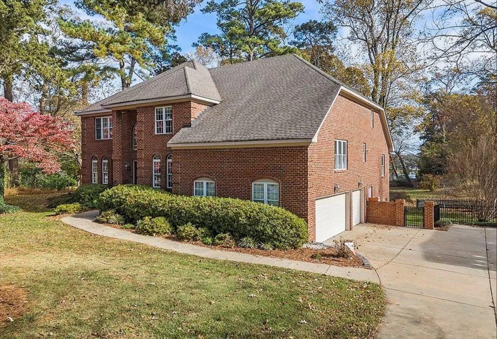 Brick suburban house with gray roof, white garage door, and driveway surrounded by trees and shrubs