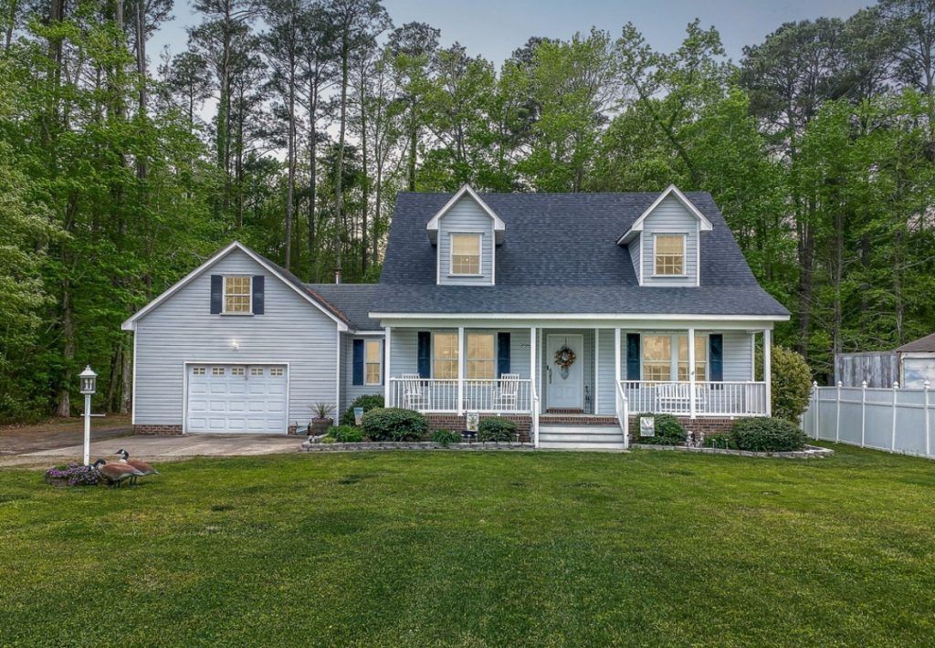 Gray house with white porch and detached garage on a grassy lot, backed by trees