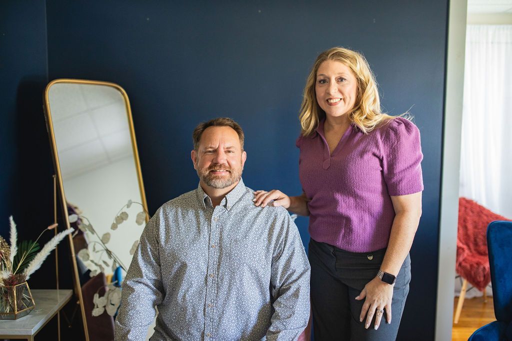 Two people posing indoors beside a gold-framed mirror, in front of a blue wall.
