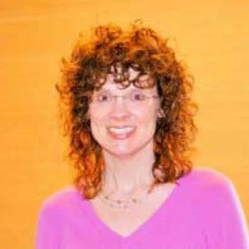 A woman with curly hair and glasses is wearing a pink shirt and smiling.