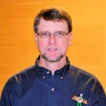 A man wearing glasses and a blue shirt is standing in front of a wooden wall.