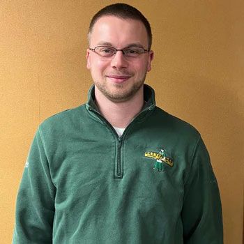 A man wearing glasses and a green sweater is standing in front of a yellow wall.