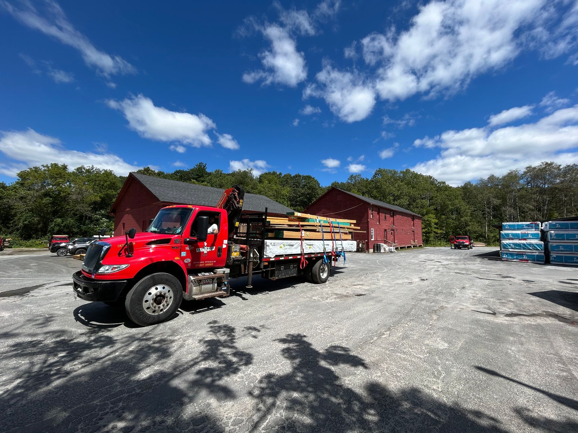 Red delivery truck loaded with lumber parked in a gravel lot near red buildings under a blue sky.