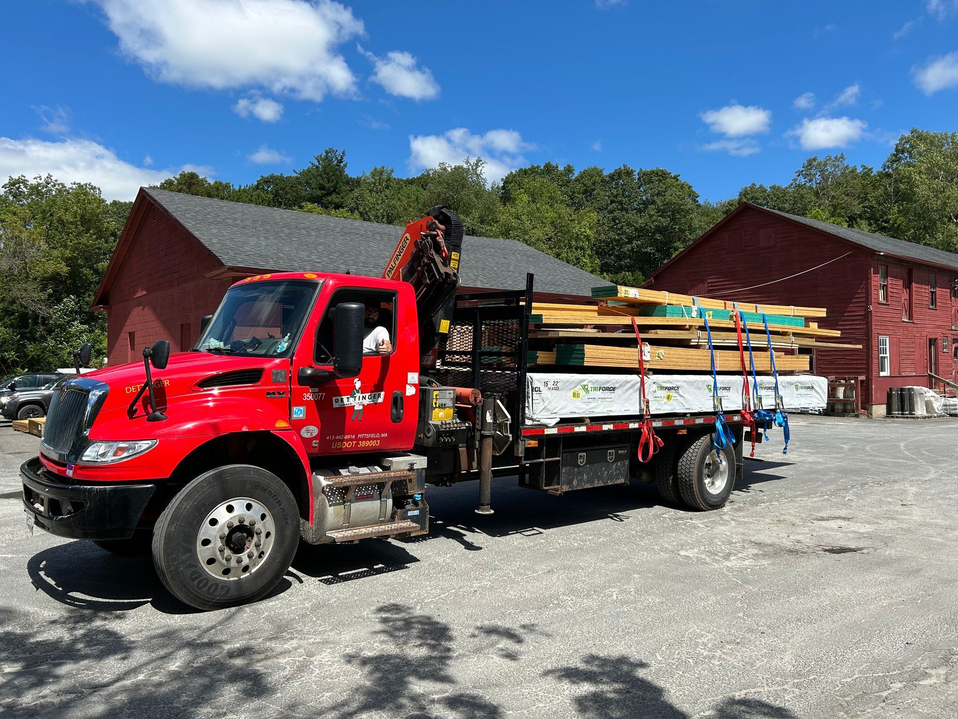 Red truck carrying lumber, parked in front of two red buildings under a blue sky with trees.