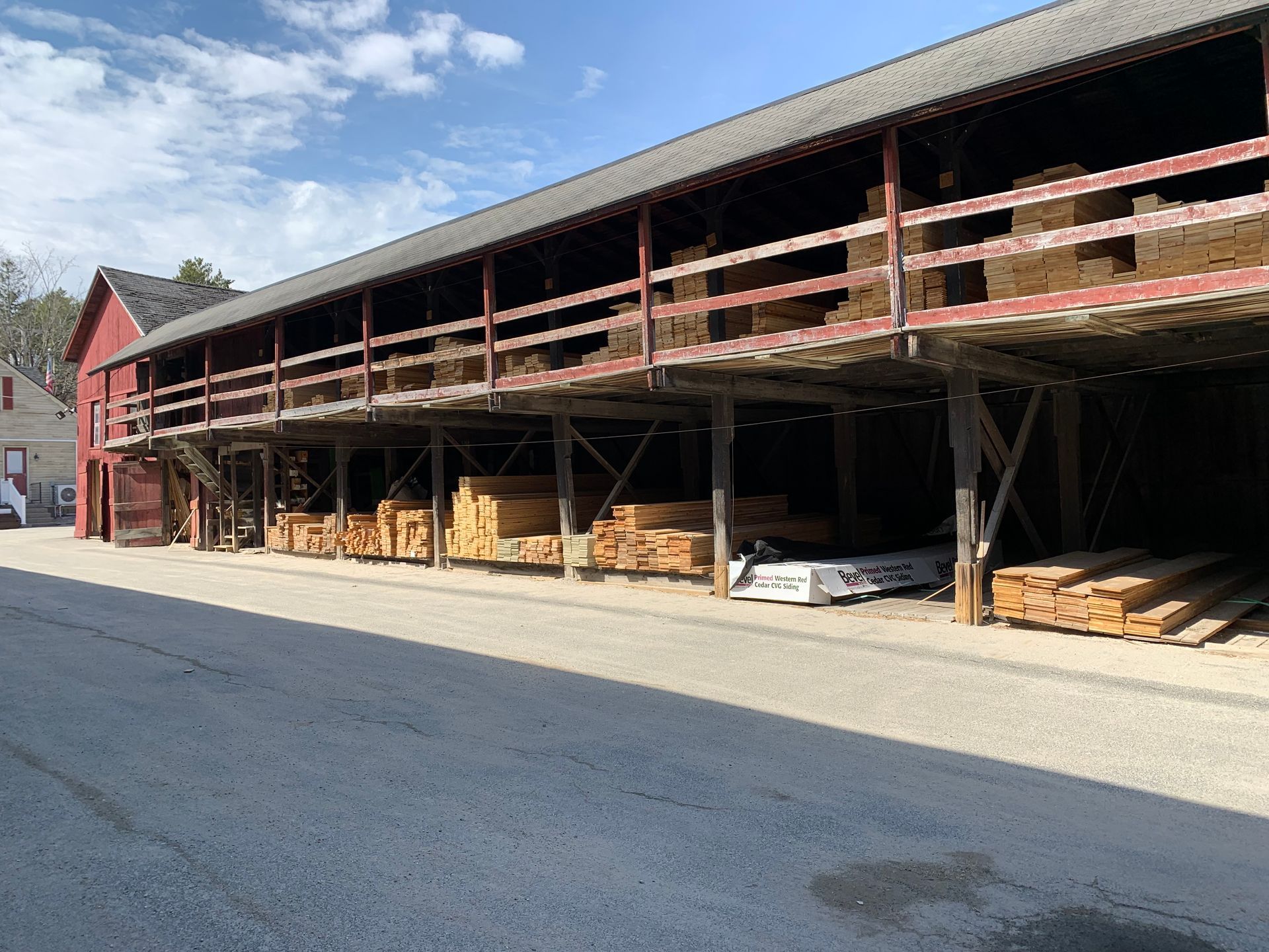 A lumber storage shed with stacked wood, red exterior, and a gravel road in front under a blue sky.