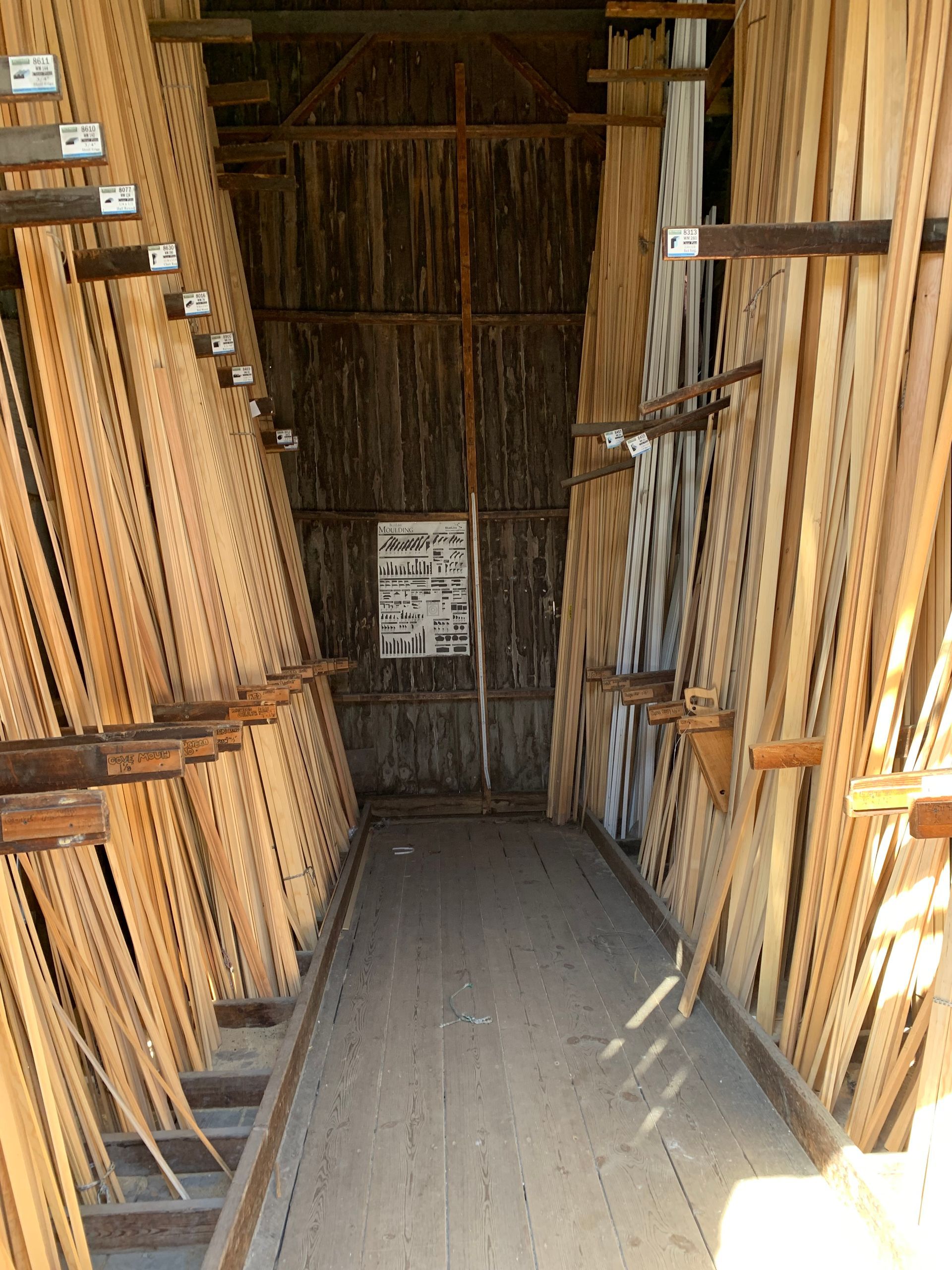 Inside a lumber storage shed, rows of long wooden boards are stacked on racks. A narrow walkway extends into the shed.