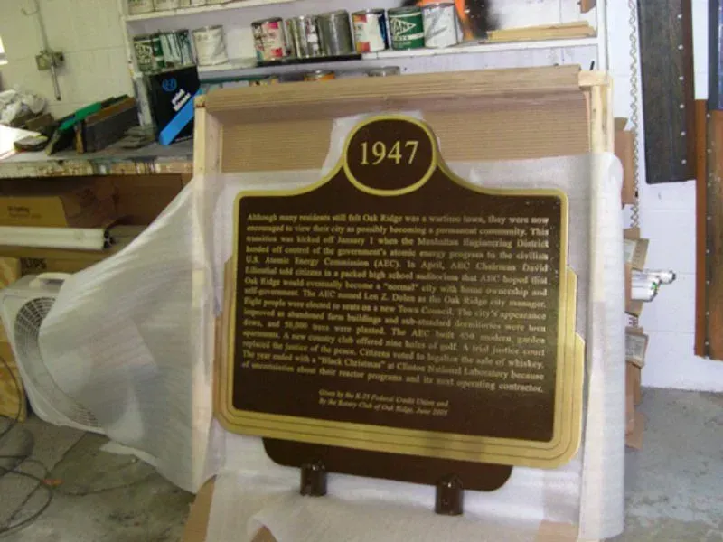 Historical marker from 1947 in a wooden shipping frame. Brown and gold, text about a city's beginnings.