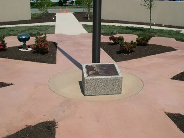 Flagpole with plaque on a concrete base, set in a plaza with flowerbeds and path.