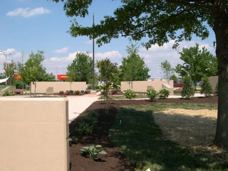 Landscaped area with beige walls, trees, mulch, and flowers under a sunny blue sky.