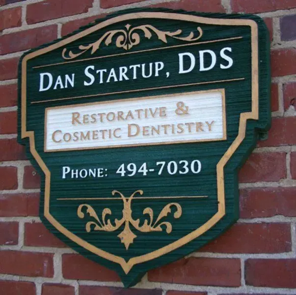Wooden sign for Dan Startup, DDS, on a brick wall, with green and gold lettering, advertising restorative and cosmetic dentistry, and a phone number.