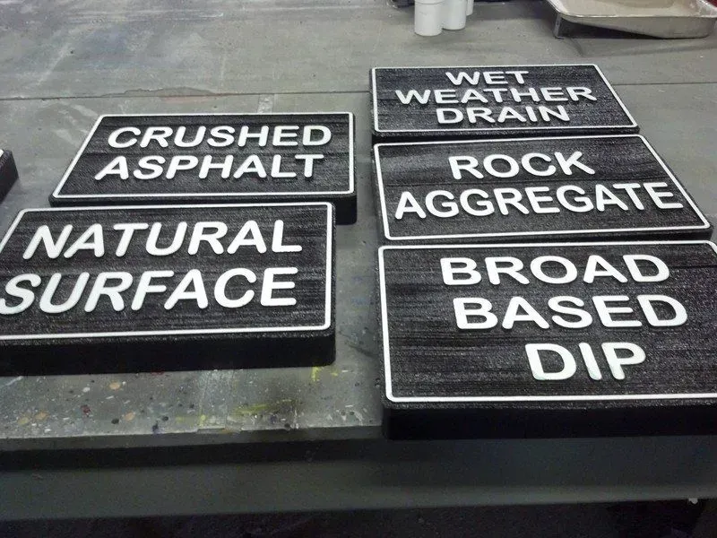 Six black rectangular signs with raised white lettering, on a gray surface. Labels include 