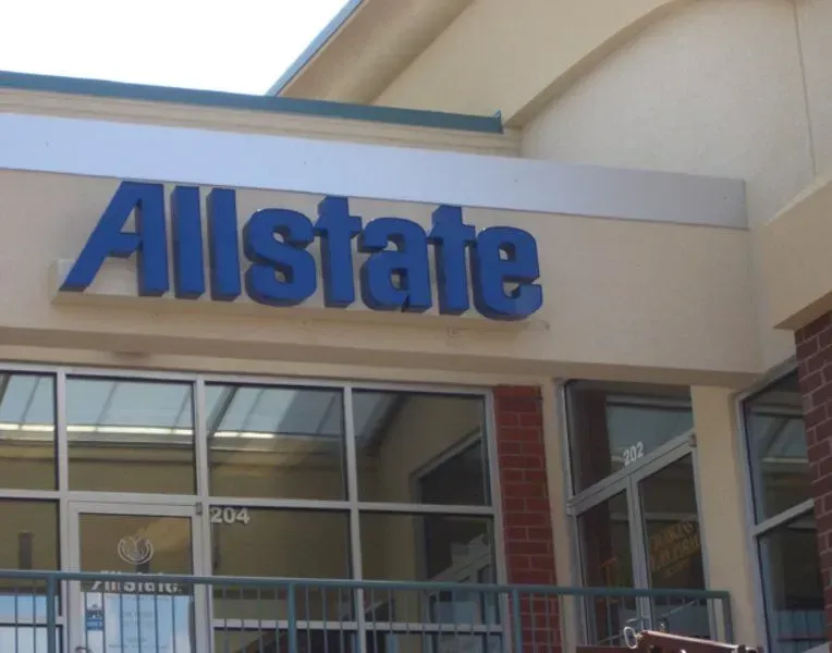 Allstate storefront with a blue sign above glass windows; beige building with a brick wall to the right.