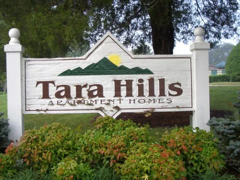 Sign for Tara Hills Apartment Homes, featuring mountain art and a sun symbol, set amidst greenery.