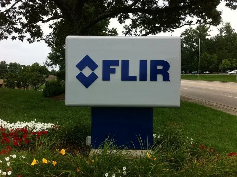 Sign for FLIR Systems, blue logo and text on a white background, in front of a green lawn and trees.